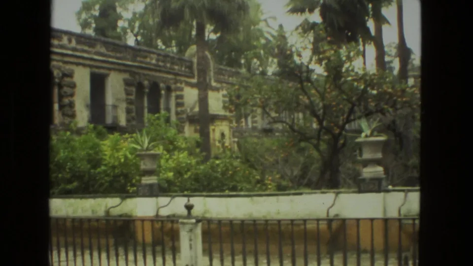 1978 Seville frame with trees, planted courtyard edge, iron fence, white wall, and historic stone facade.