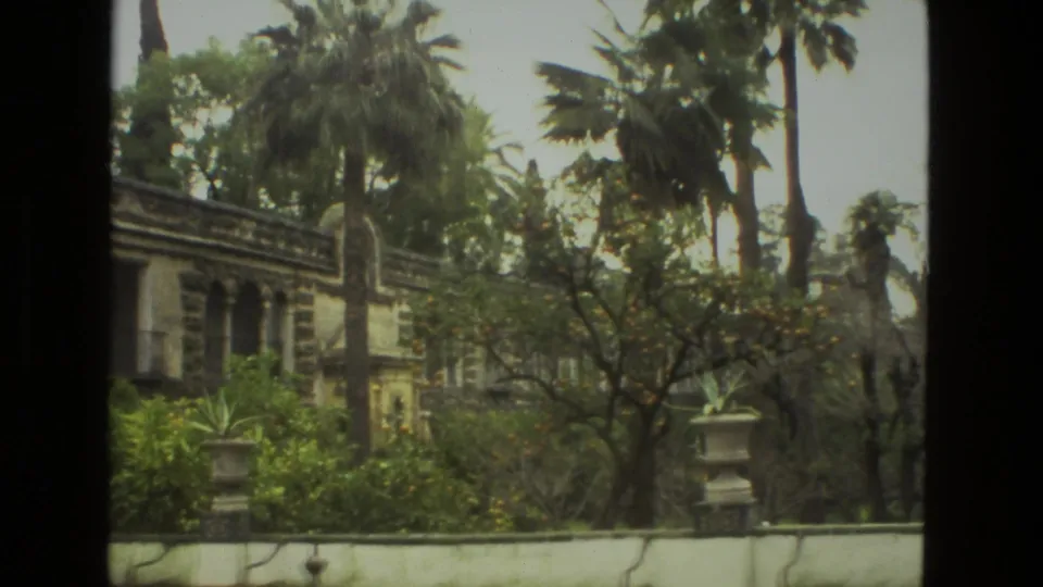 1978 Seville home-movie frame showing an older masonry structure partly obscured by palms, hedges, and dense garden planting.