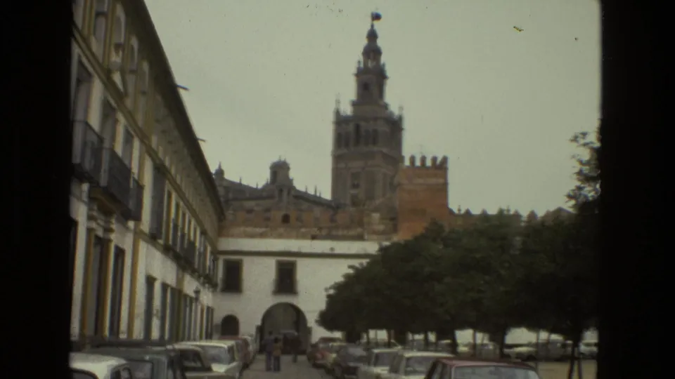 The 1978 Seville home-movie frame shows the cathedral tower beyond a street lined with parked cars, trees, and small distant figures near an archway.