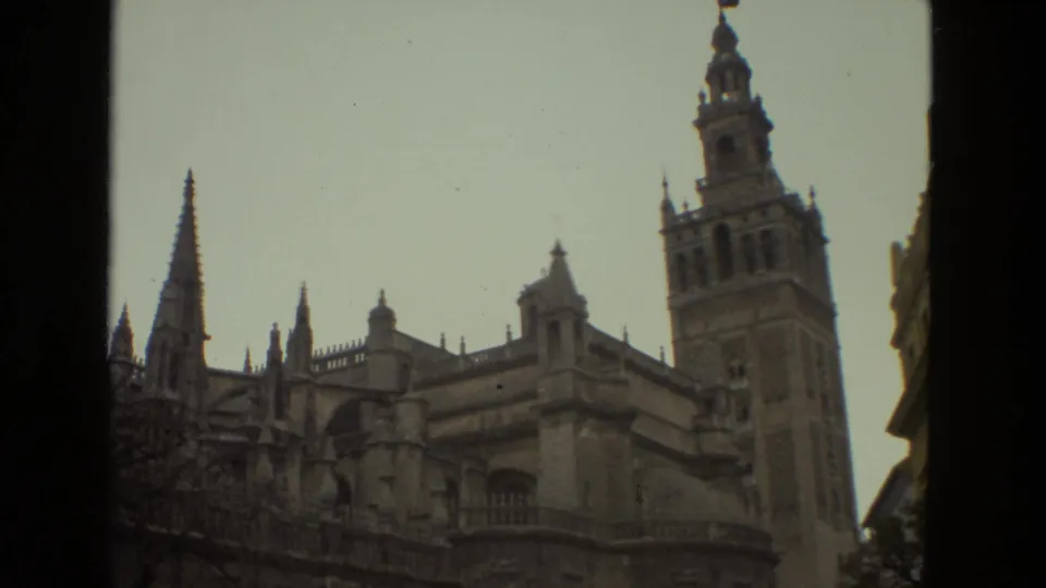1978 home-movie frame looking up at Seville Cathedral, with Gothic spires and the Giralda tower prominent against the sky.
