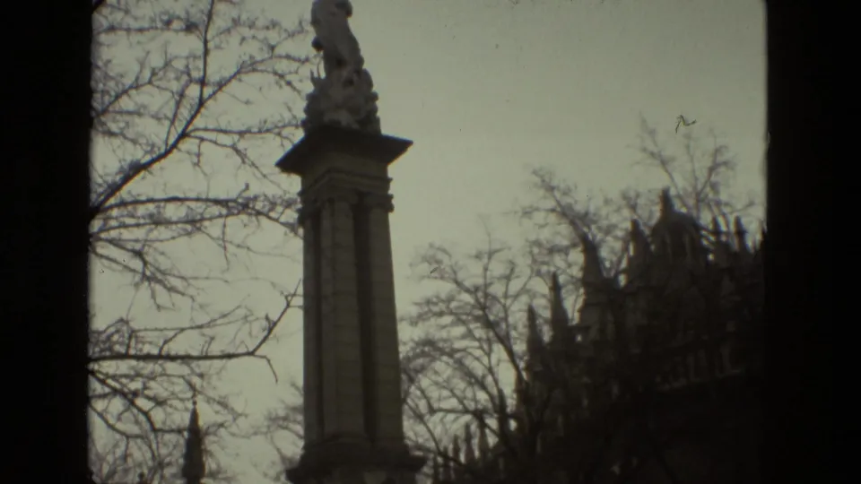 Handheld 8mm view in Seville showing a tall monument column, bare branches, and the cathedral partly obscured in the background.