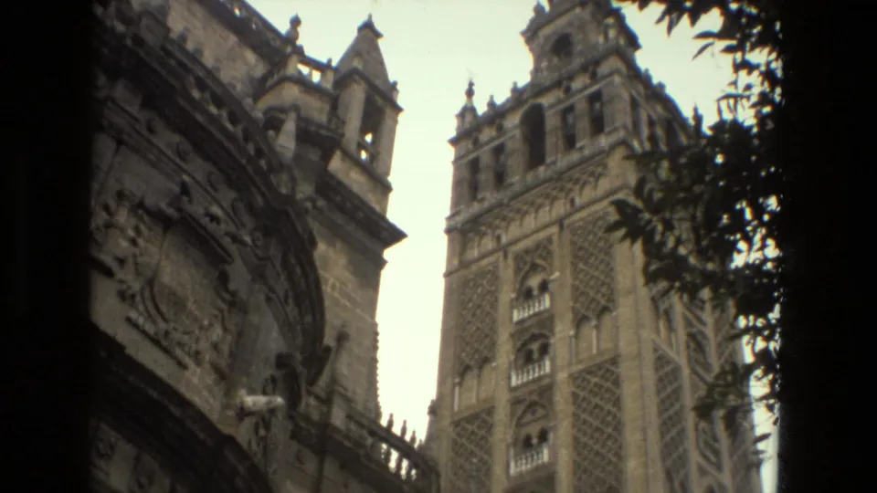 1978 Seville home-movie view of the Giralda tower behind carved facade details and nearby foliage.