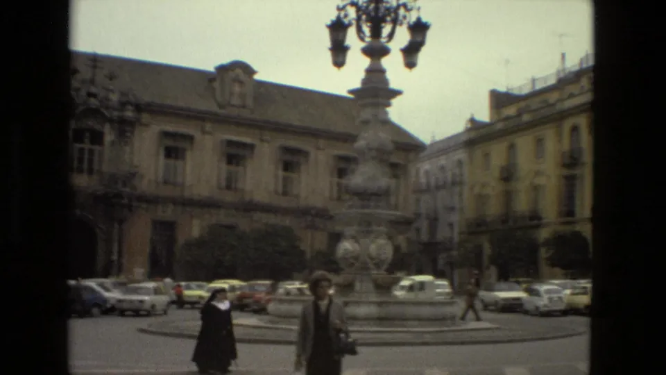 A nun and another pedestrian move through a Seville square in 1978 near a stone fountain, cars, and historic buildings.