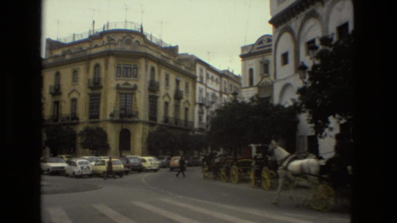 Wide street corner with historic buildings, parked cars, pedestrians, and horse-drawn carriages.