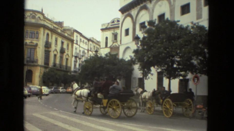 Horse-drawn carriages with passengers move along a Seville street in 1978, with trees, parked cars, and historic facades behind them.