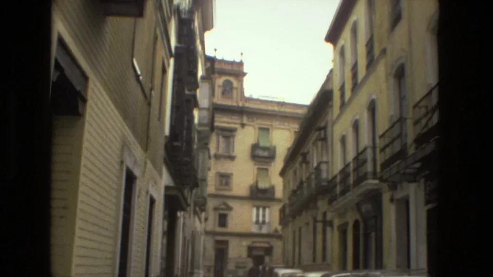 Narrow Seville street in 1978 with tall stucco buildings, wrought-iron balconies, a parked car, and a historic facade ahead.
