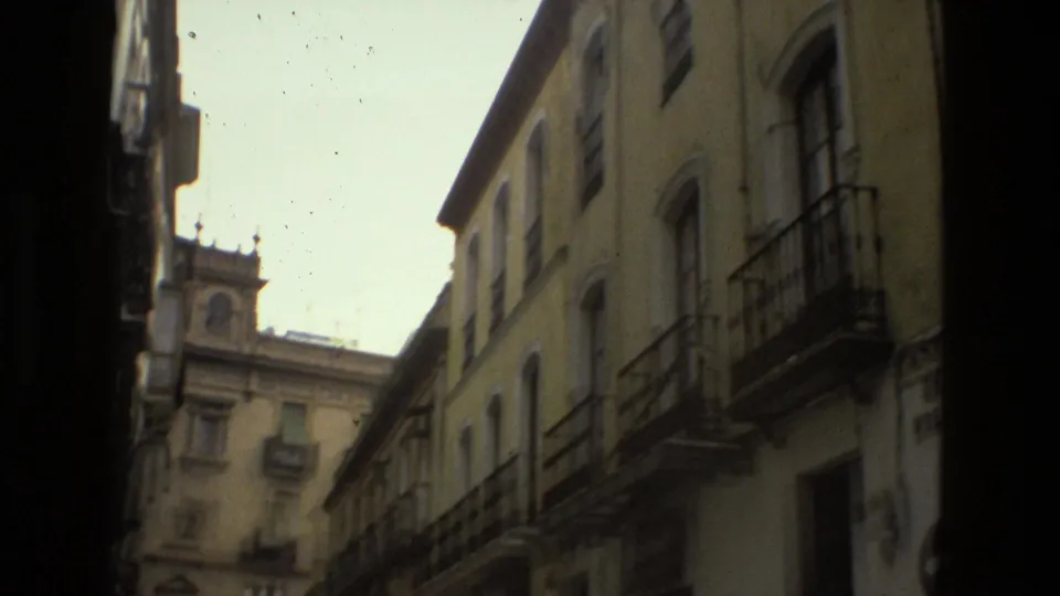 Handheld 8mm view down a narrow Seville street in 1978, framed by tall buildings, balconies, windows, and a distant facade.
