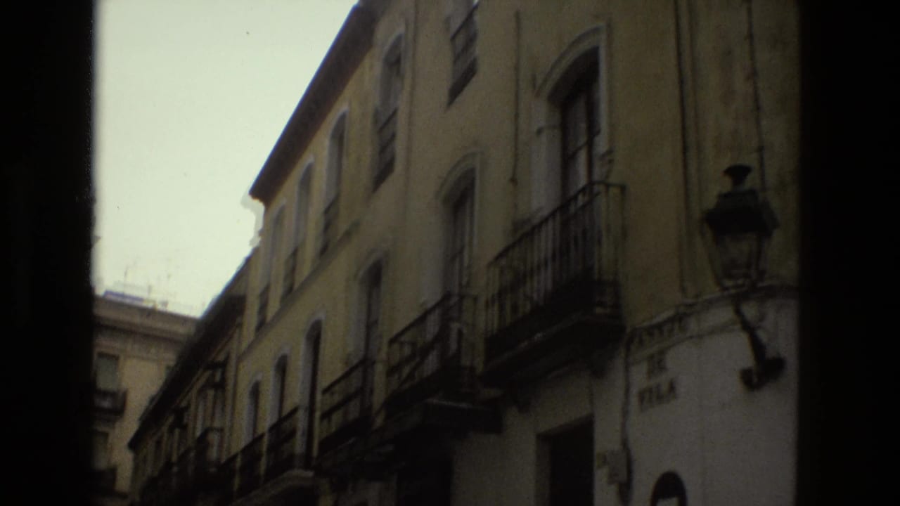 1978 Seville home-movie frame looking upward along a narrow street lined with stucco facades, wrought-iron balconies, and a street lamp.