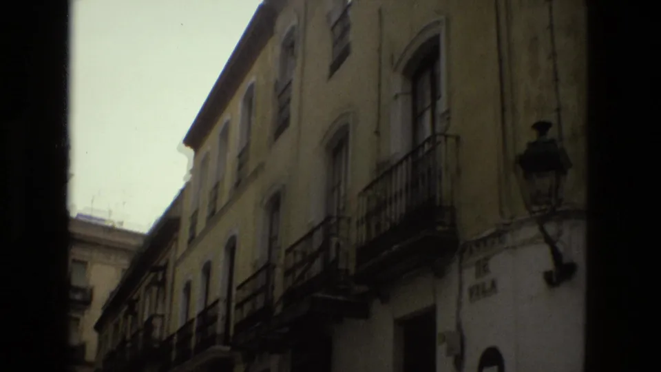 1978 Seville home-movie frame looking upward along a narrow street lined with stucco facades, wrought-iron balconies, and a street lamp.