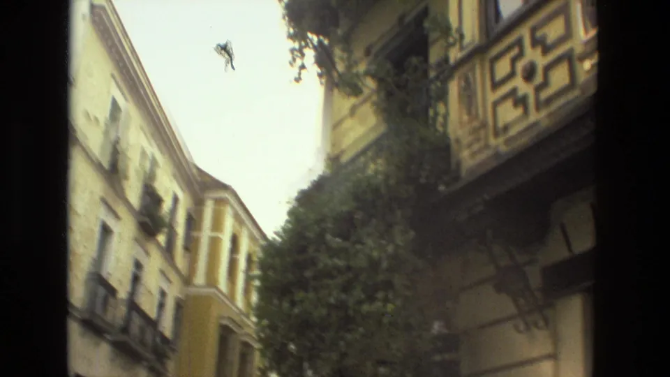 1978 Seville frame looking through a narrow historic street with stucco buildings, decorative balconies, a leafy tree, and open sky.