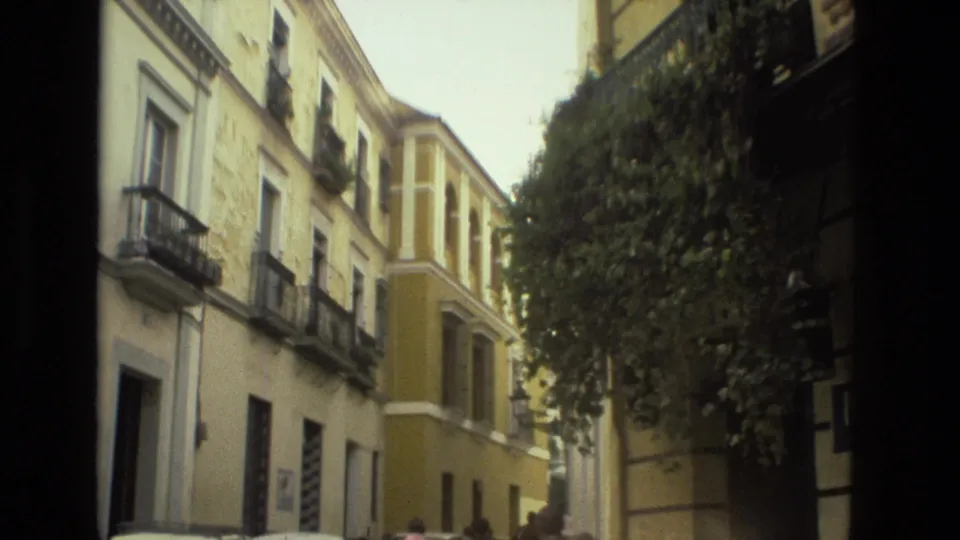 1978 Seville street scene with pale buildings, yellow corner facade, balconies, leafy trees, and passersby partly visible.