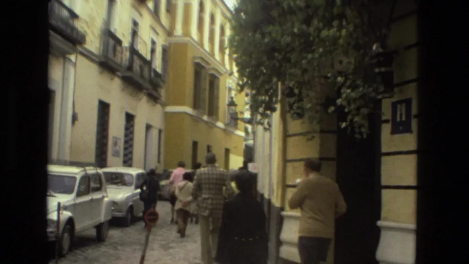 1978 8mm street view in Seville with pedestrians walking away from camera, parked compact cars, balconies, and cobblestone paving.