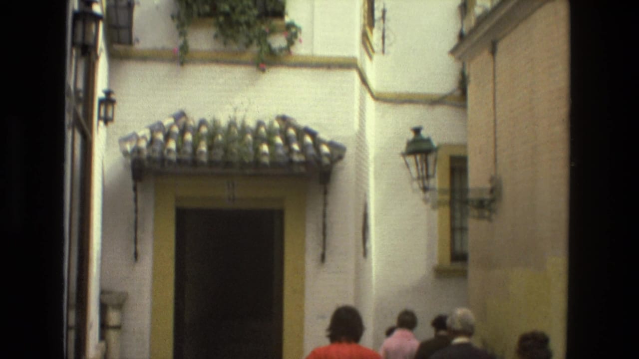 1978 Seville home-movie frame showing visitors in a shaded alley approaching a whitewashed facade with a tiled canopy and doorway.