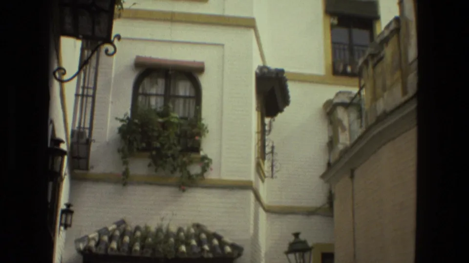 1978 Seville home-movie frame looking up at a white facade with arched window, balcony plants, ironwork, and lanterns.