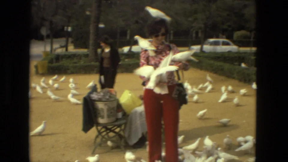 A woman in red pants and a patterned top holds white pigeons in a Madrid square, with more birds spread across the ground.