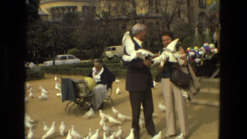 An older man in a dark suit and a woman in light trousers hold white pigeons in the Madrid square as birds gather at their feet.