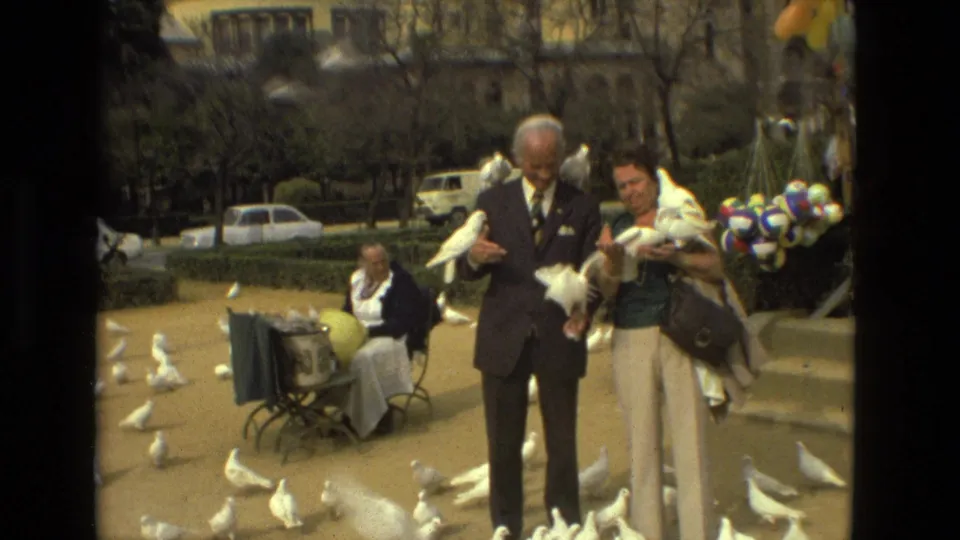 A man in a dark suit and a woman in light trousers hold white pigeons in a Madrid courtyard while more birds gather around them.