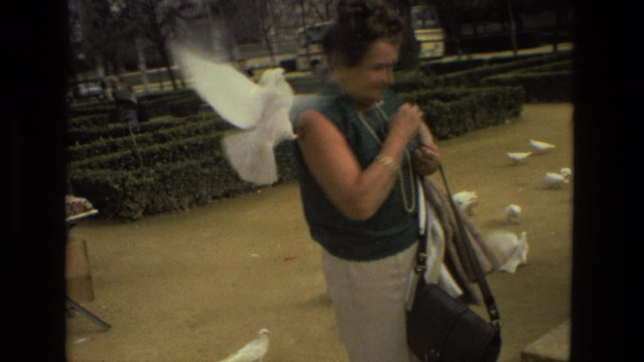 In 1978 Madrid home-movie footage, a woman with a handbag stands near white doves on a sandy path in a hedged park.