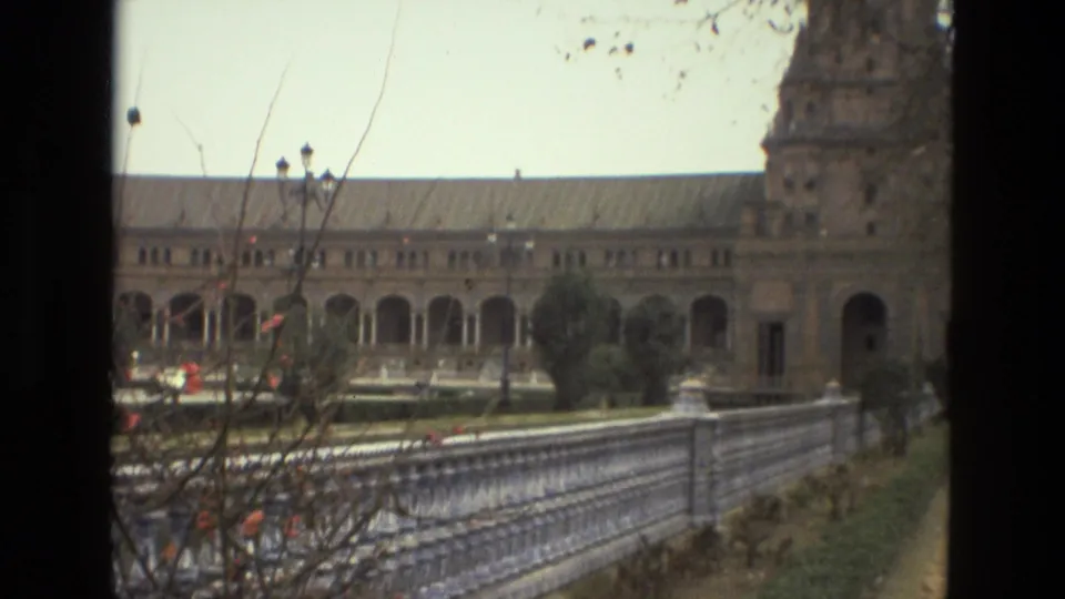 1978 Madrid home-movie view of a long colonnade and tower edge seen past branches and a tiled balustrade.
