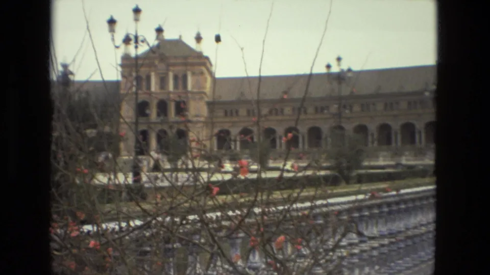 1978 Madrid 8mm frame with the palace's central tower and arched wings seen past branches, lampposts, and a stone railing.