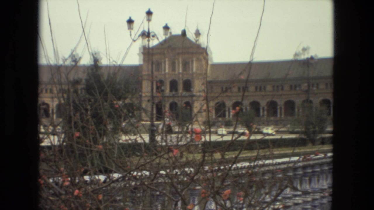 1978 8mm travel frame of Plaza de España in Seville, viewed through branches toward arched architecture and plaza paving.