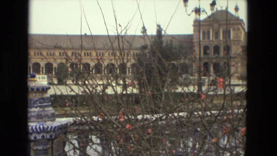 Visitor-shot 8mm view across a tiled fountain and bare branches toward Plaza de España architecture in Seville.