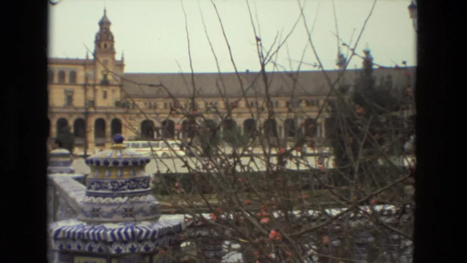 Late-1970s 8mm frame of a tiled fountain and arched Plaza de España buildings seen past winter branches.