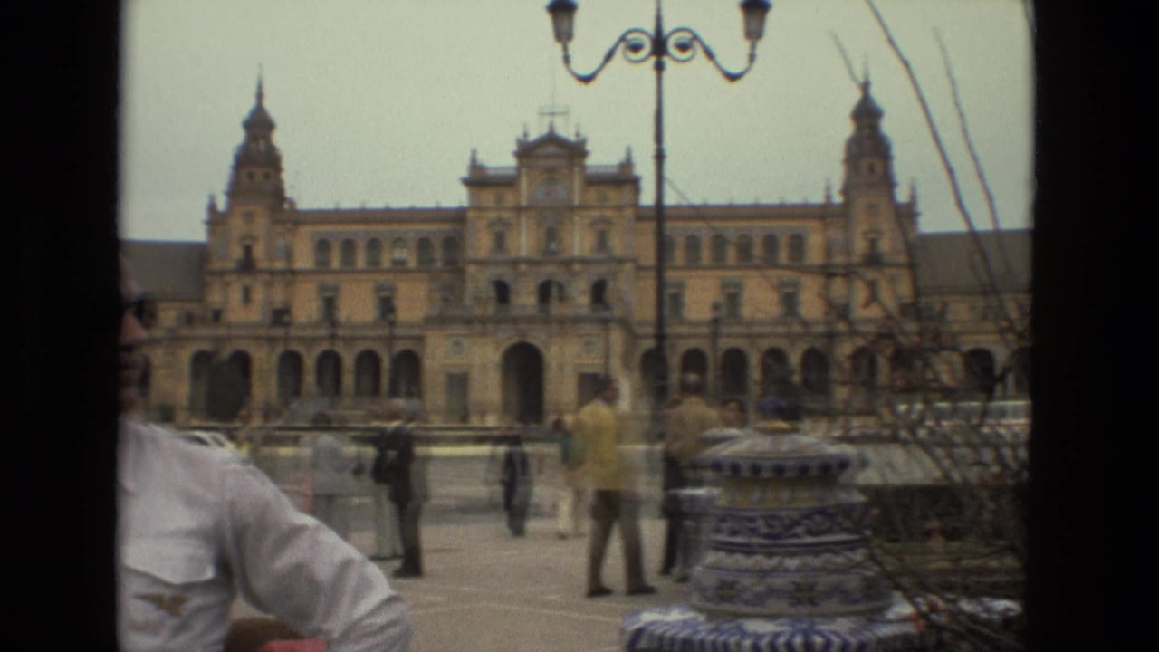 1978 Madrid home-movie frame showing a grand arched building, pedestrians in the plaza, street lamps, and a partial man in white in the foreground.