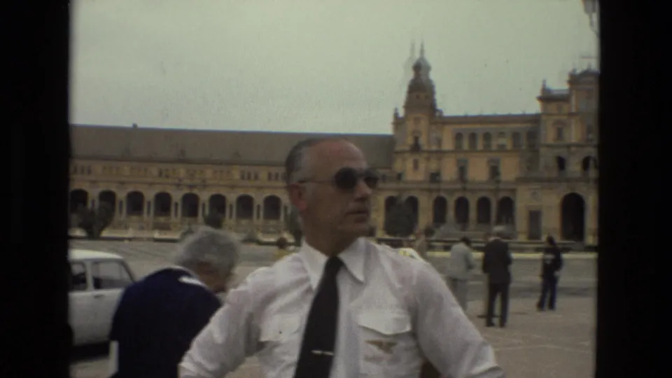 1978 Madrid frame with a man in a white uniform and sunglasses in the foreground, pedestrians, cars, and an ornate plaza building behind him.