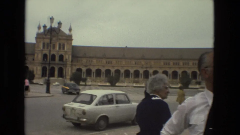 1978 Madrid home-movie frame showing a compact car, pedestrians, a partial foreground figure, and a long arched building across the plaza.