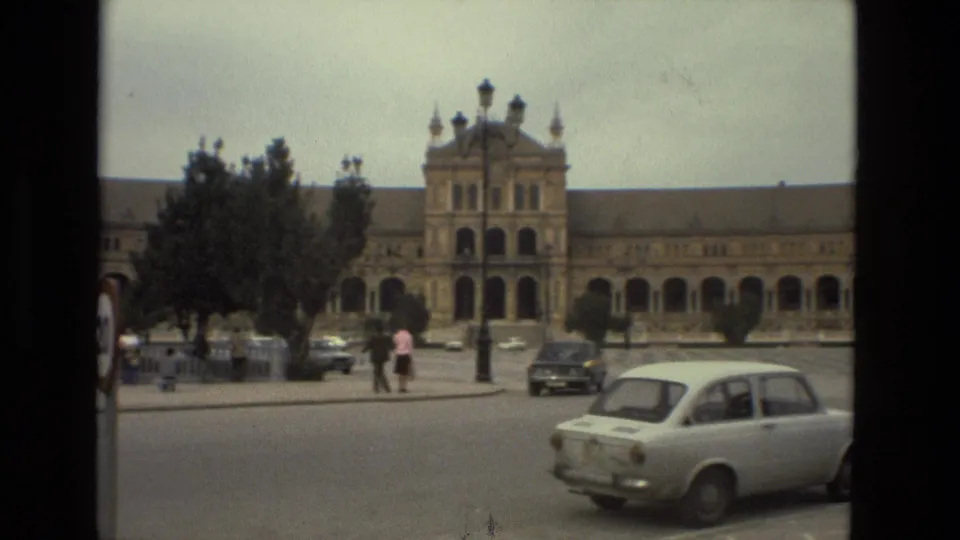 1978 Madrid frame showing a wider view of the plaza with compact cars, scattered pedestrians, trees, lamps, and ornate arched architecture.