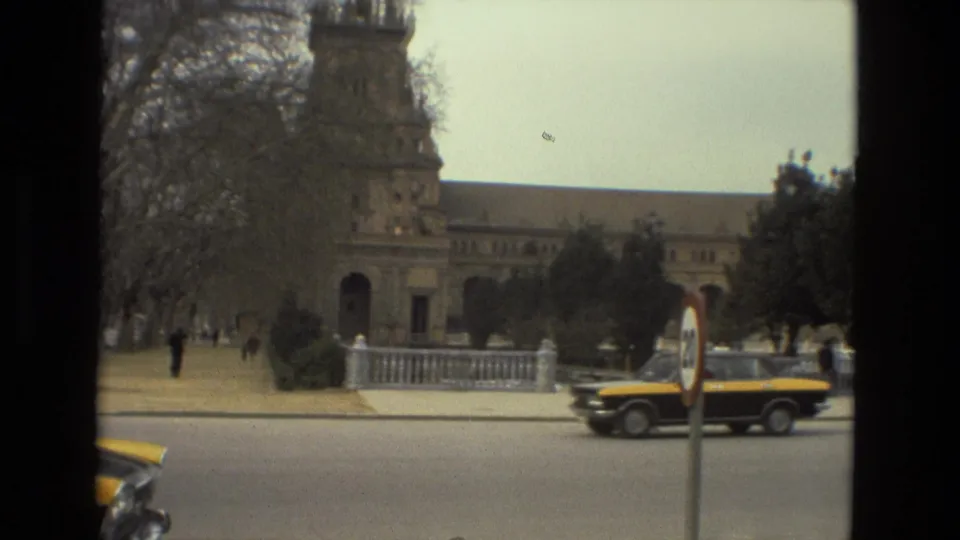 Black-and-yellow car on a Madrid street near a roundabout sign and towered historic building, with pedestrians in the distance.