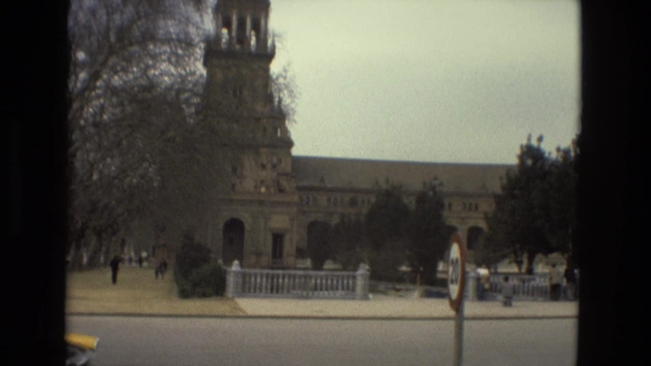 1978 Madrid home-movie frame showing a large historic building across a road with trees, signposts, and small pedestrian figures.