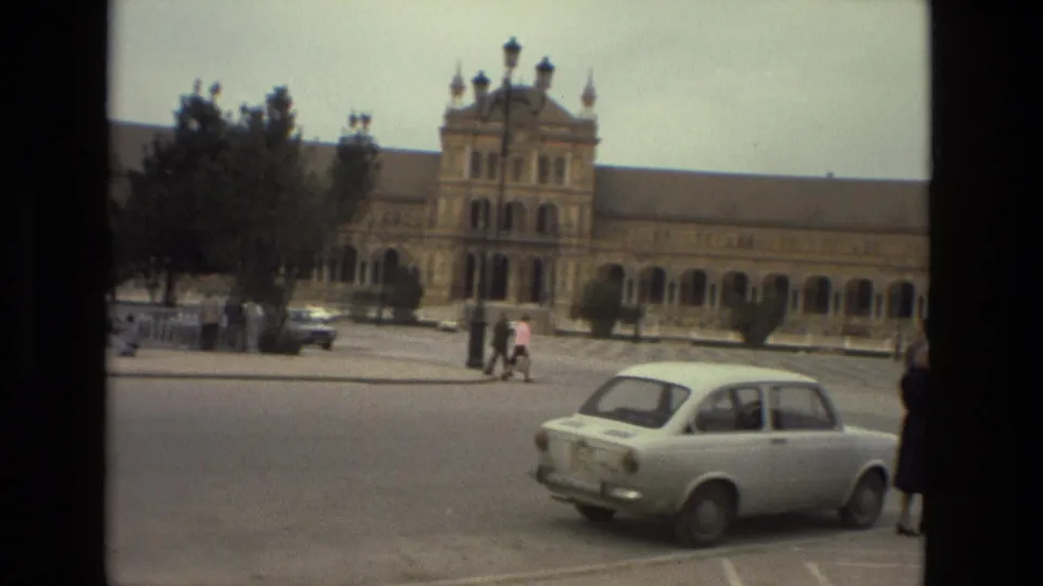 1978 Madrid street view with a white compact car, pedestrians, trees, and a long arched historic building in the background.