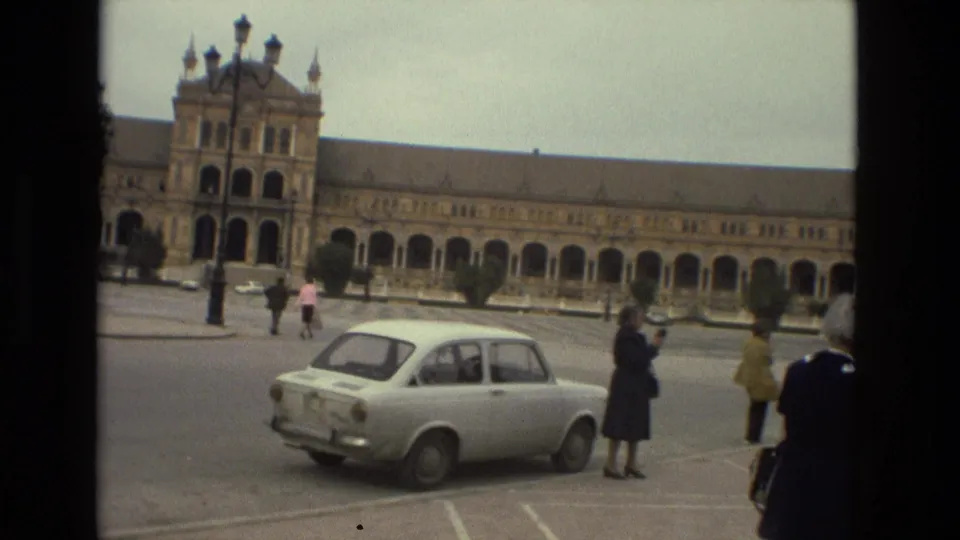 1978 Madrid home-movie frame showing a white compact car and pedestrians before a long colonnaded historic facade.
