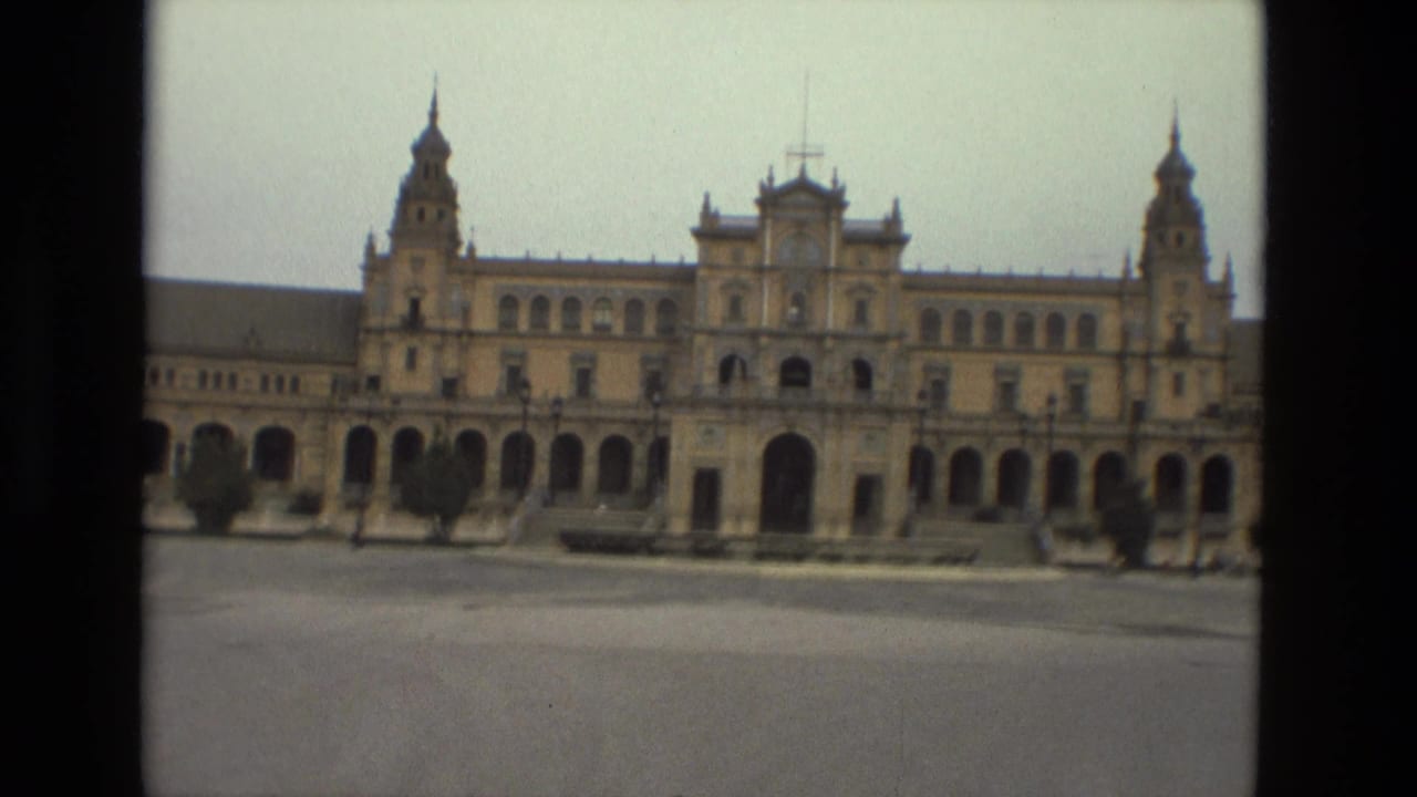 Ornate colonnaded building with towers across a wide paved plaza.
