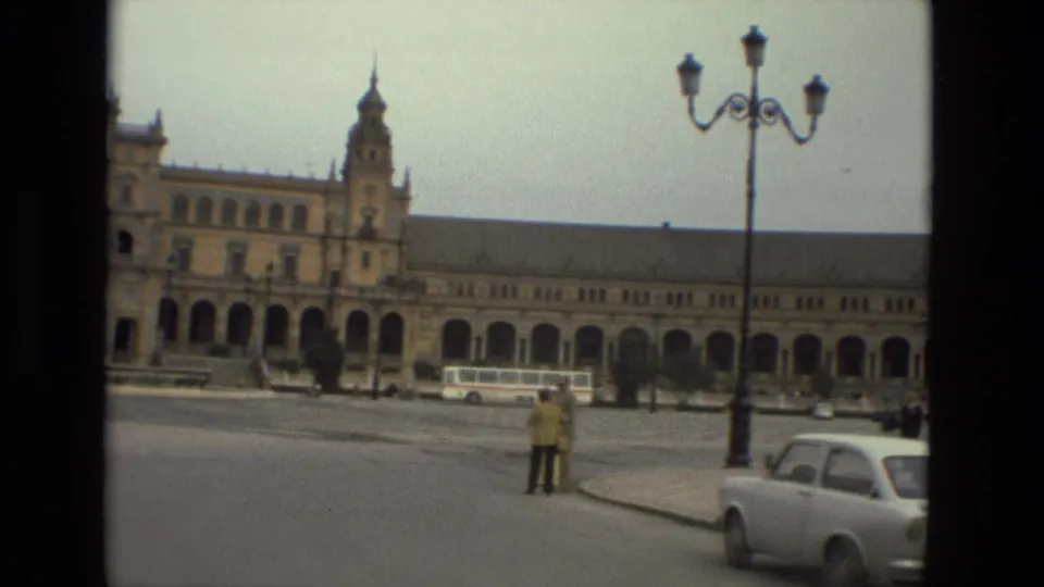 1978 plaza scene with the camera panning past a streetlamp, bus, white compact car, and two pedestrians near the curb.