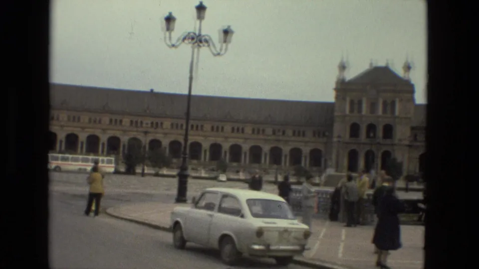 End frame showing a white compact car, groups of pedestrians, a streetlamp, and the long arched facade of the plaza building.