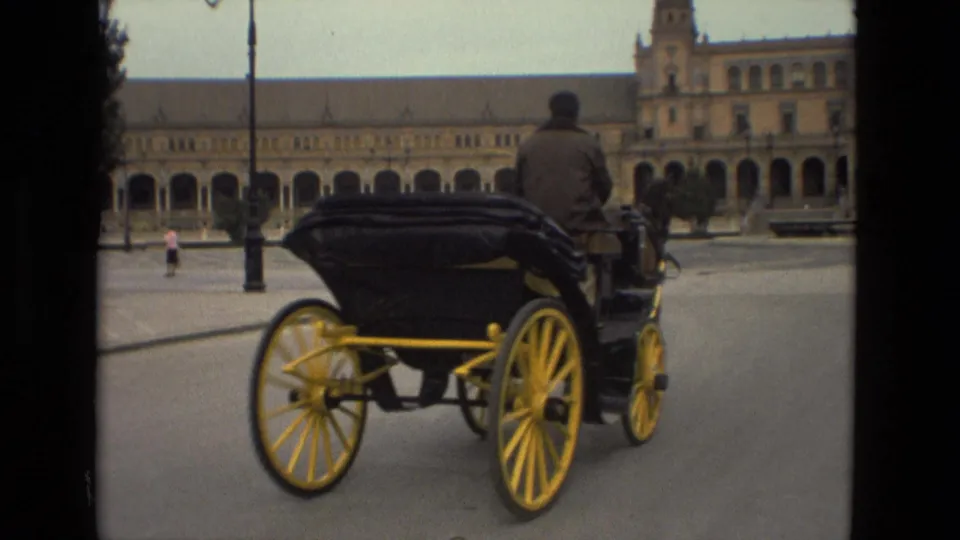 A horse-drawn carriage moves through Plaza de España in Madrid, with historic architecture and street lamps behind it.
