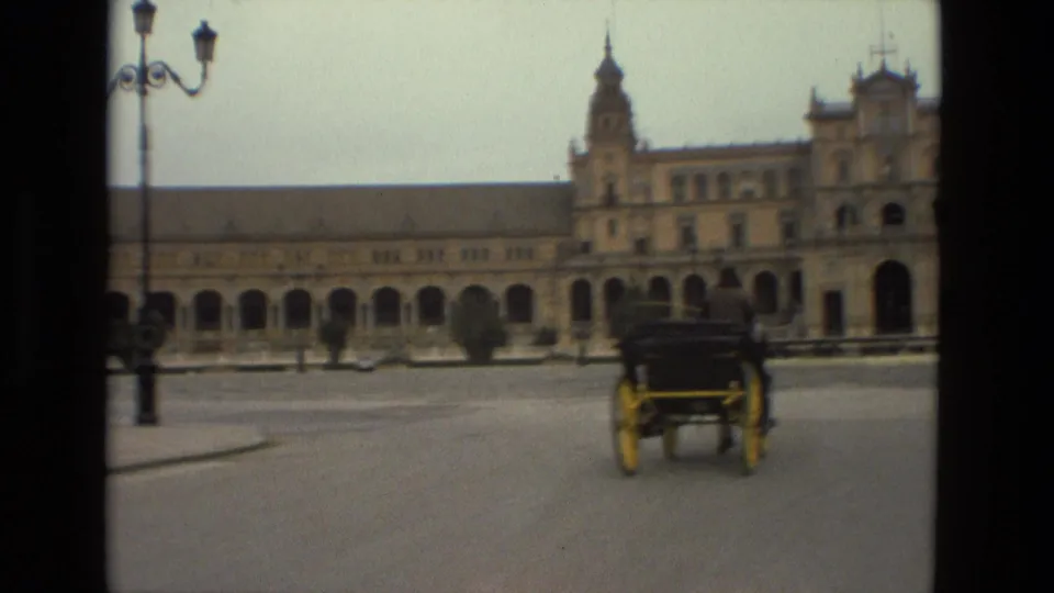 A distant carriage crosses the open Madrid plaza, with lamps, trees, and the arched facade visible through soft 8mm focus.