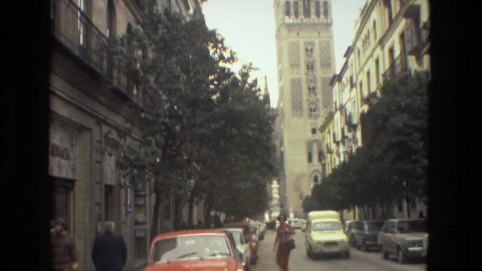 A Madrid street view shows pedestrians, parked cars, a yellow taxi, and a tall tower rising beyond tree-lined sidewalks.