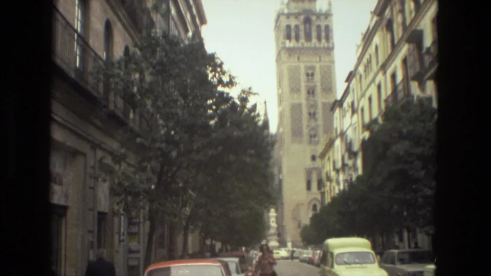 1978 Spain home-movie frame of a tall historic tower rising beyond trees, balconies, parked cars, and a narrow city street.