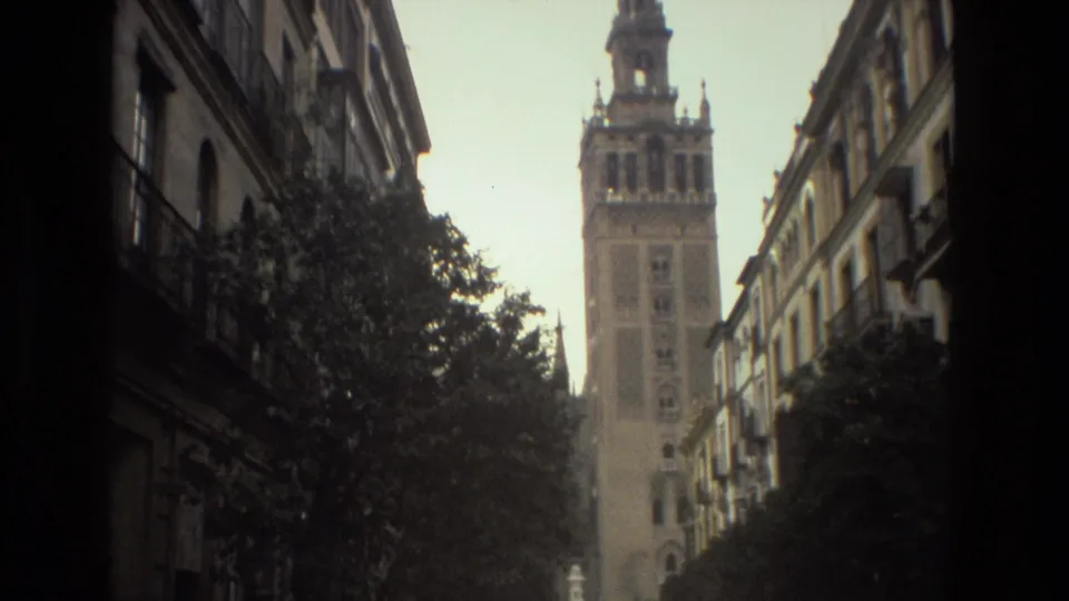 1978 Spain street view looking toward a tall tower, with shadowed facades, trees, and parked vehicles in the foreground.