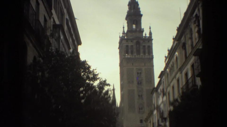 1978 Spain home-movie view of the tower from a narrow street, with dark trees, apartment balconies, and parked cars below.