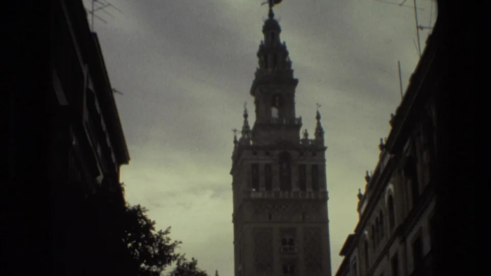 1978 Spain street frame showing the tower against a cloudy sky, with dark building edges and tree silhouettes in the foreground.