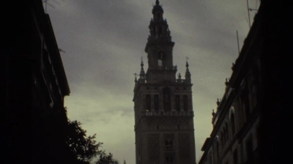 1978 8mm travel frame showing a tall historic tower above shadowed street buildings in Spain, with cloudy sky behind it.