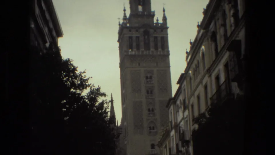 A shadowed 1978 street view in Spain shows a historic tower beyond narrow buildings and trees, filmed from a handheld 8mm perspective.