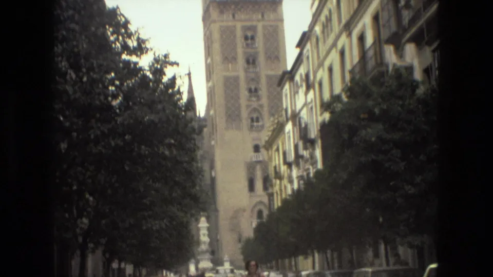 The 8mm frame brightens to show a tree-lined Spanish city street with parked cars, buildings, and the tower in the distance.