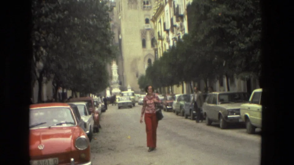 A woman walks along a 1978 Spanish city street with parked compact cars, trees, buildings, and the historic tower behind her.