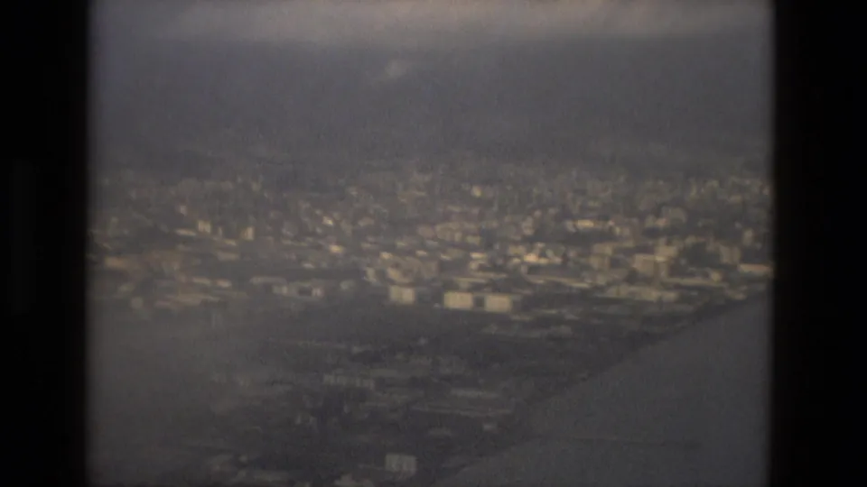 Home-movie aerial frame over Barcelona in 1978, with muted water, city blocks, and distant hills seen from the plane.